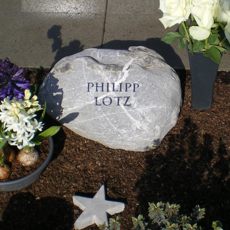 Weiß marmorierter, grauer Grabstein-Findling für ein Urnengrab,  mit schwarzen Knubbeln und eingehauener Kapitalis-Inschrift