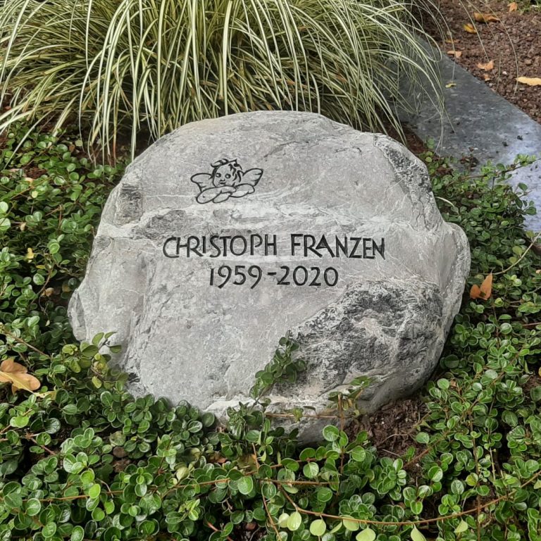 Weiß marmorierter, grauer Grabstein-Findling für ein Urnengrab,  mit schwarzen Knubbeln, Engel-Ornament à la Raffael und  eingehauener Grotesk-Inschrift