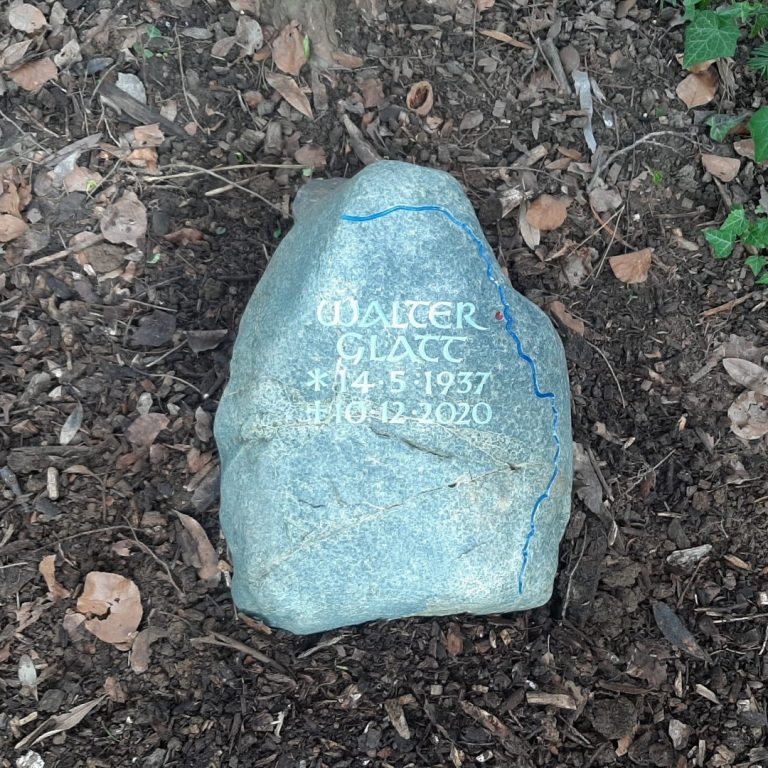 Grüner Grabstein-Findling mit eingearbeiteter Unziale-Schrift und  dem Verlauf des Rheins, sowie einem roten Punkt zur  Kennzeichnung der Stadt Köln