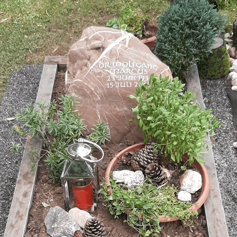 Grau-weiß-braun marmorierter Findling-Grabstein für ein Einzelgrab mit eingehauener, getönter Unzialis-Beschriftung und Grabumrandung aus Bergischer Grauwacke
